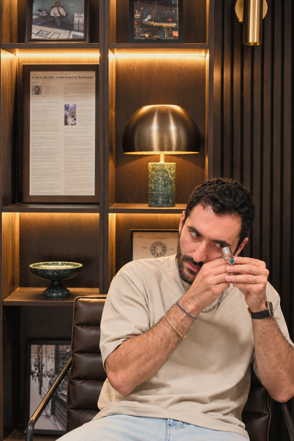 Avi examining a green gemstone through a loupe, seated in the showroom office beneath a framed article about his work