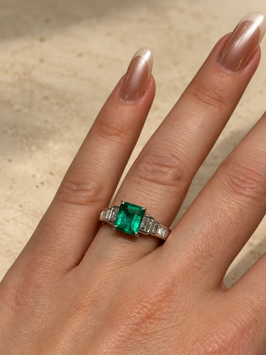 An emerald-cut emerald engagement ring in white metal, with step-cut diamond shoulders, worn on a hand