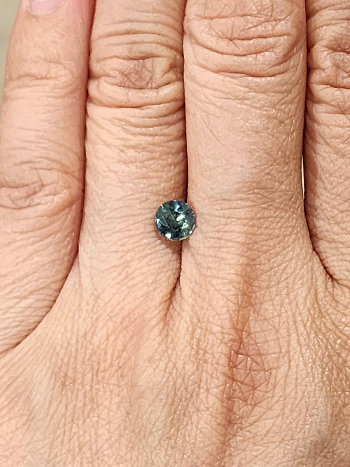 Small round green-blue sapphire gemstone shown on a finger for scale