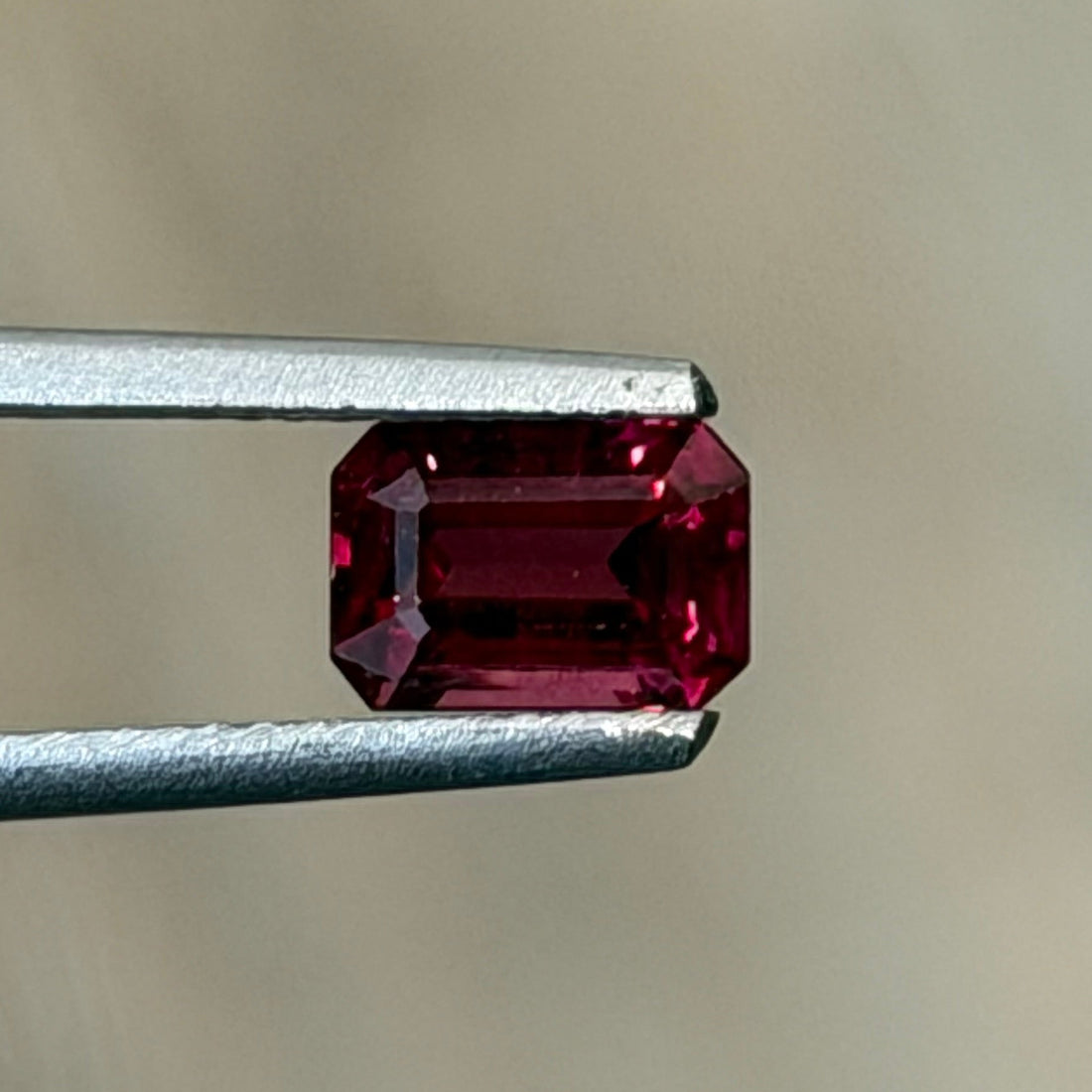 A small rectangular red gemstone held between metal tweezers against a soft beige background.