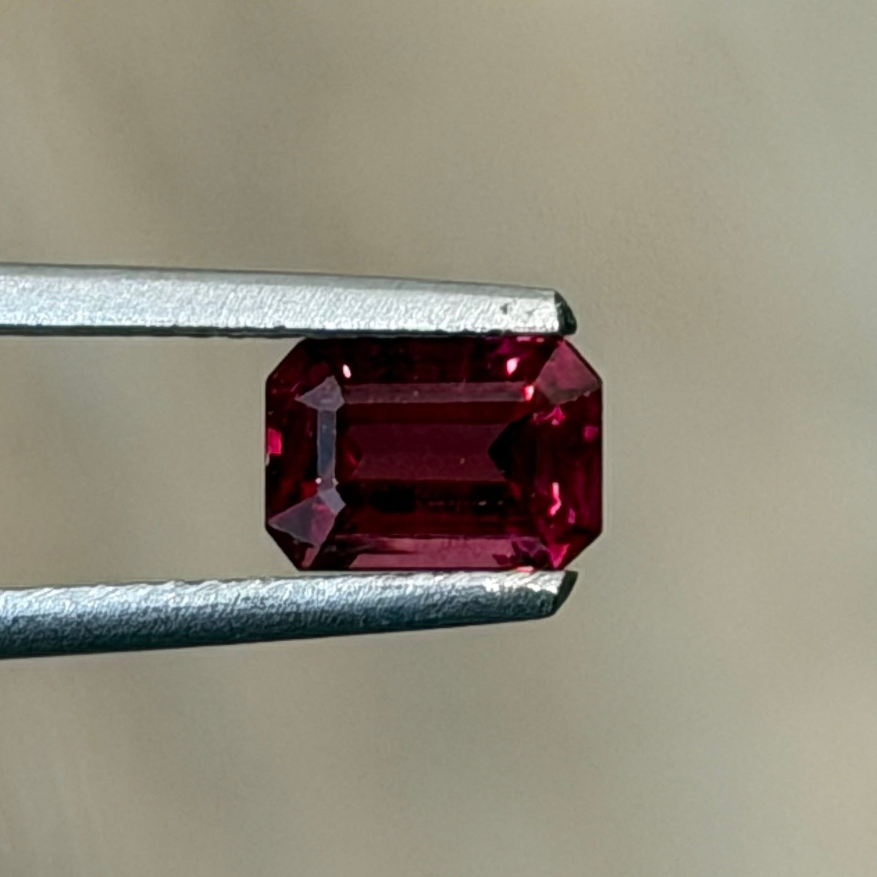 A small rectangular red gemstone held between metal tweezers against a soft beige background.