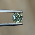 A small round-cut light green gemstone held between metal tweezers against a soft beige background.
