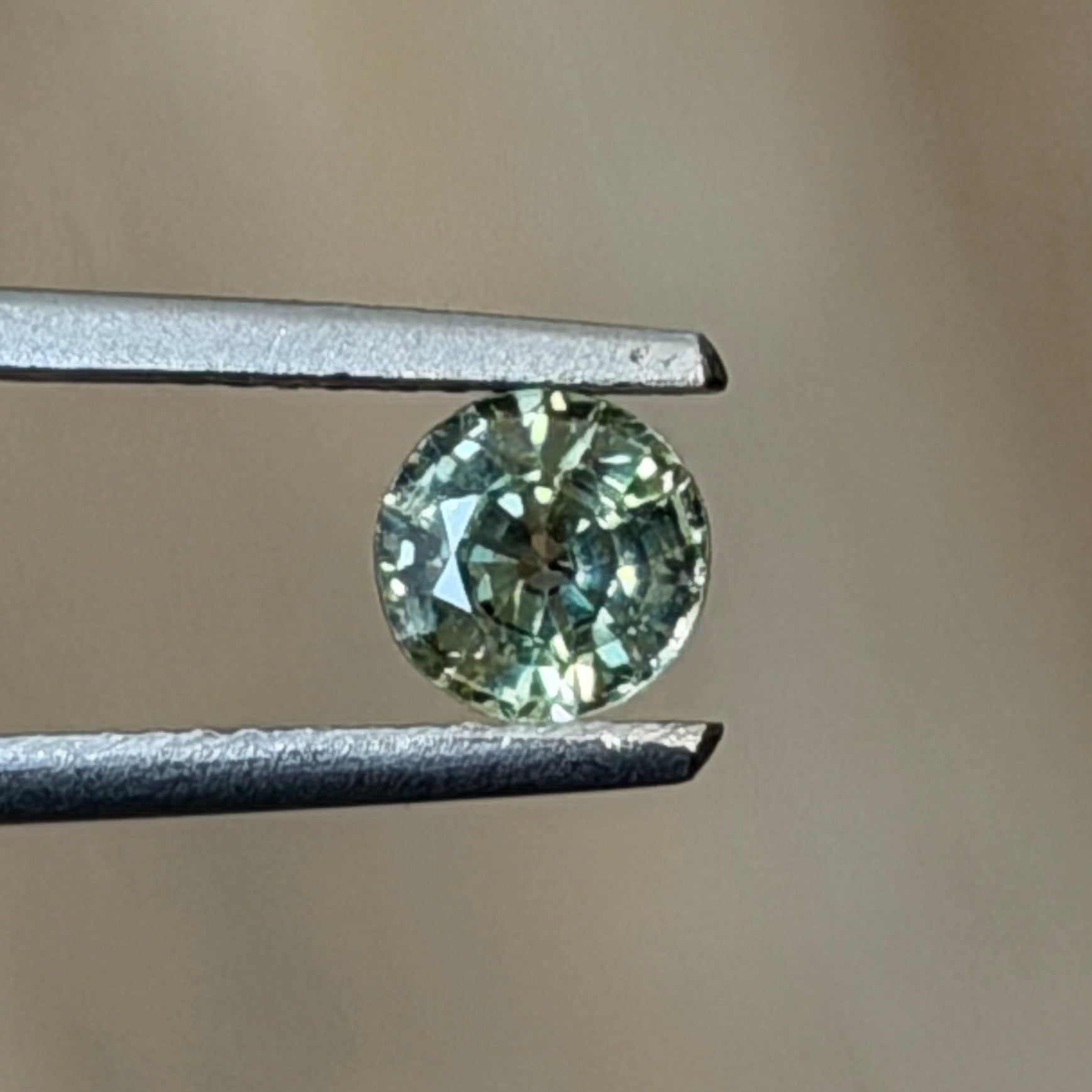 A small round-cut light green gemstone held between metal tweezers against a soft beige background.