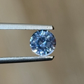 A round brilliant-cut light blue gemstone held in silver metal tweezers against a soft, blurred beige background.
