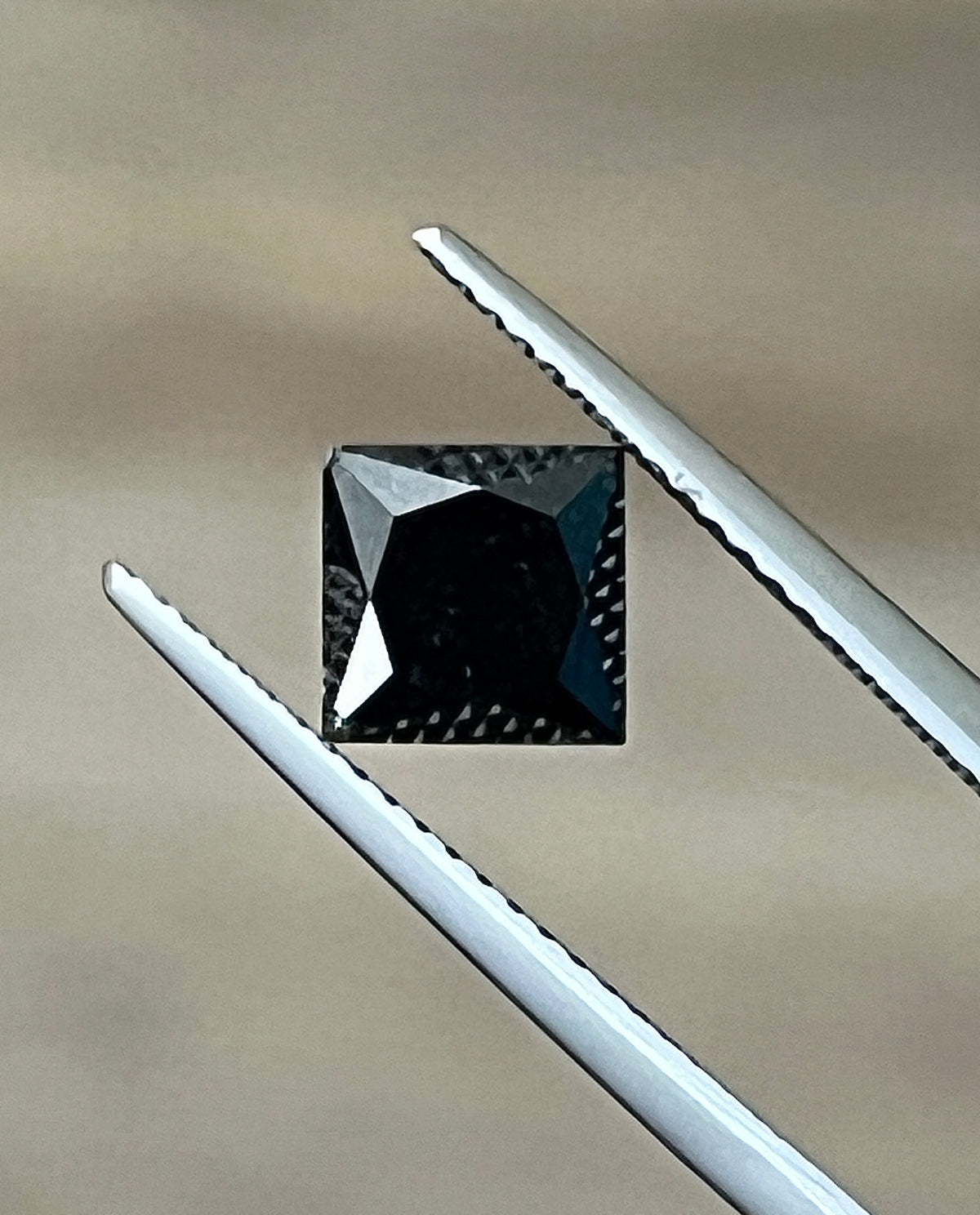 Black cushion-cut diamond held by metal tweezers, visible facets on top