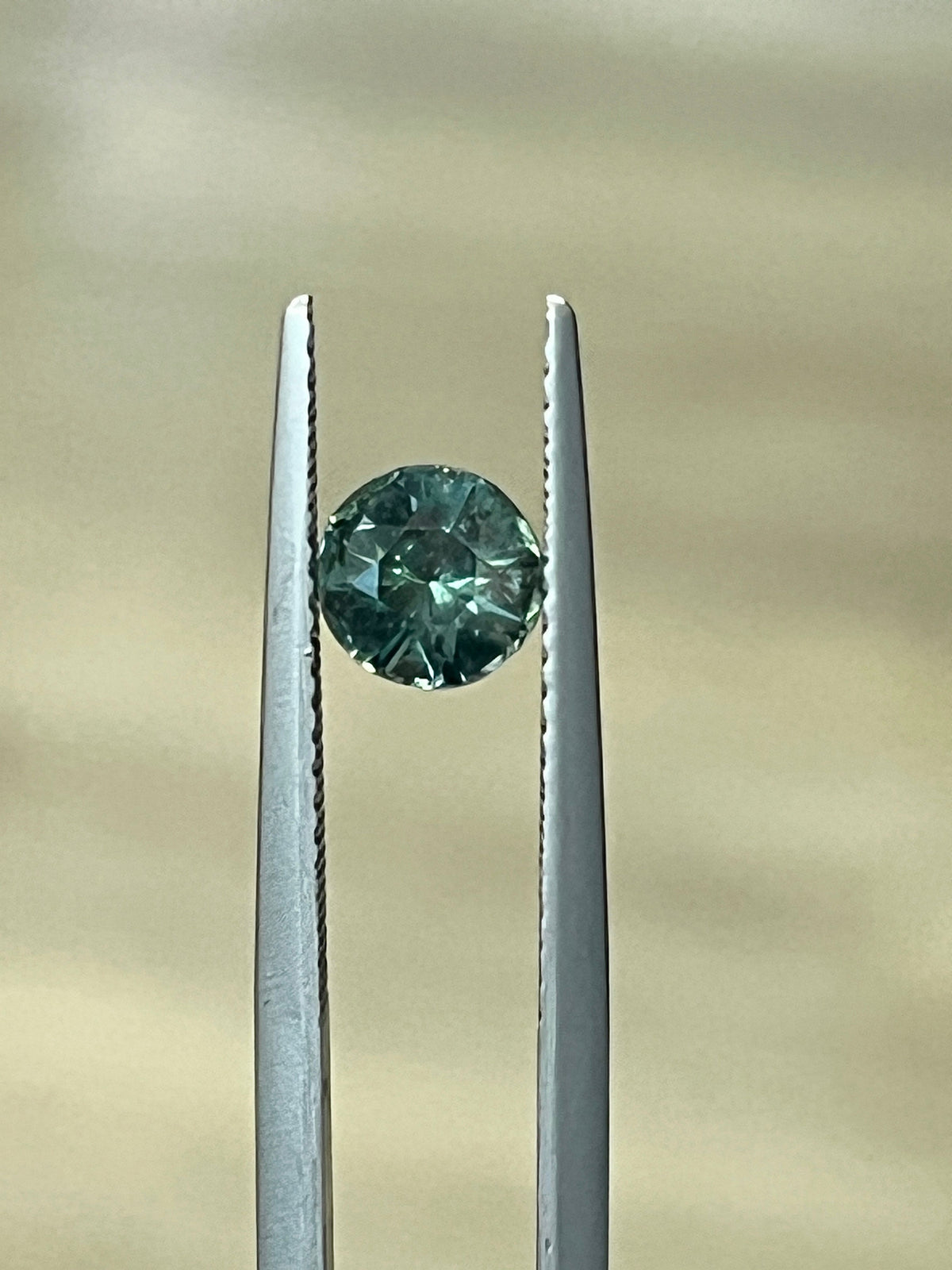 Round faceted green-blue sapphire gemstone held in tweezers