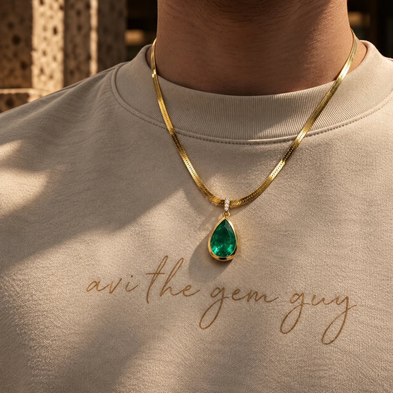 Pear-cut emerald pendant bezel-set in gold on a herringbone chain, worn over a cream Avi The Gem Guy t-shirt