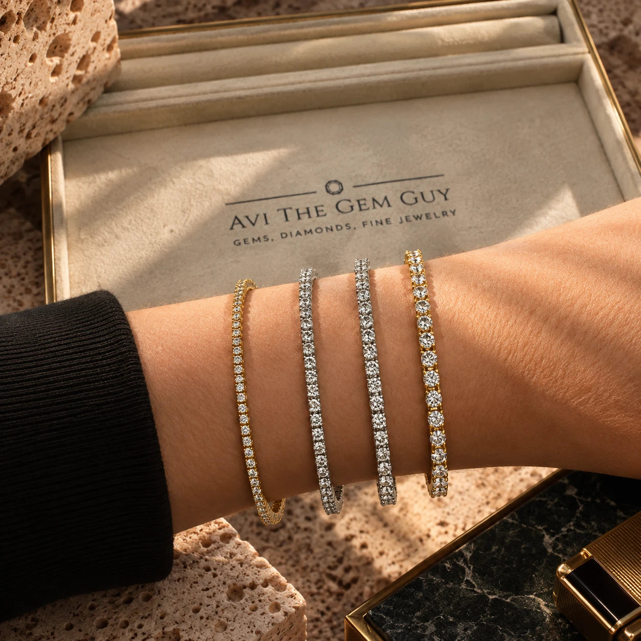 Four diamond tennis bracelets stacked on a wrist, beside an Avi The Gem Guy jewellery box