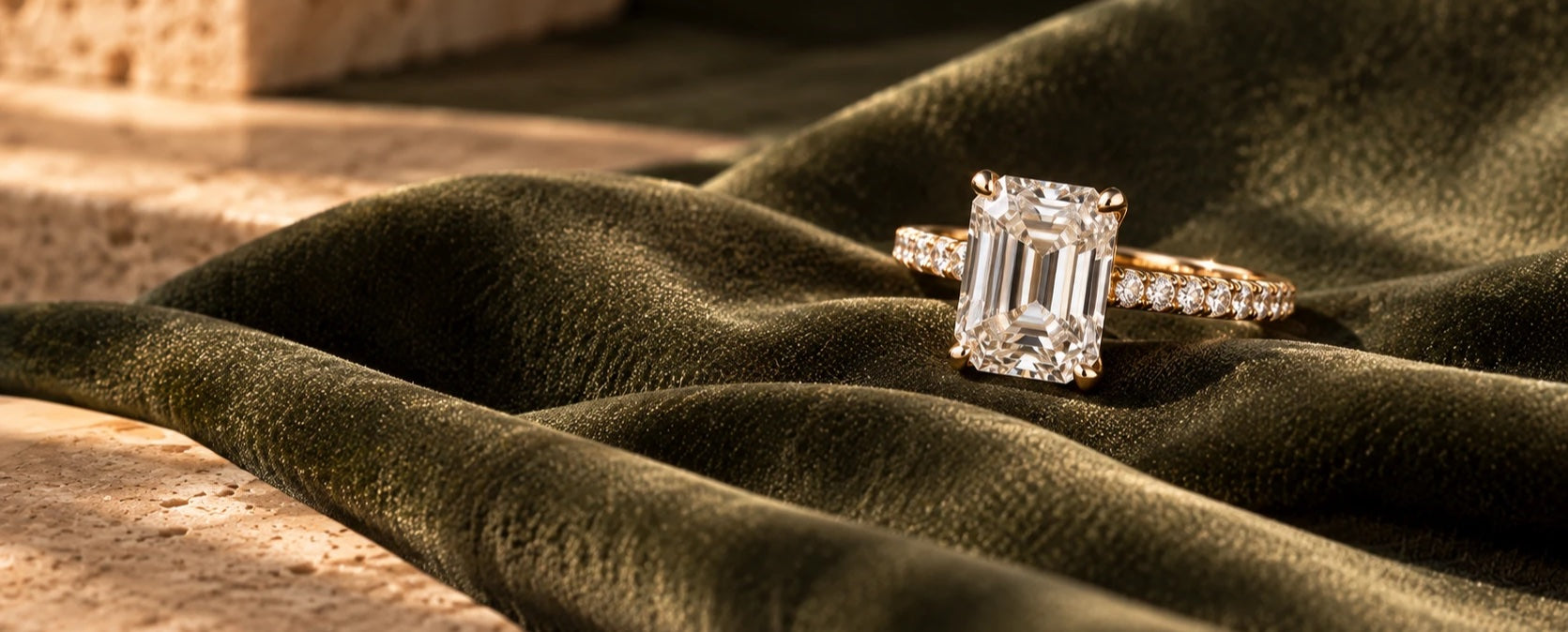 Emerald-cut diamond solitaire in yellow gold with a pave band, resting on olive velvet beside a travertine block